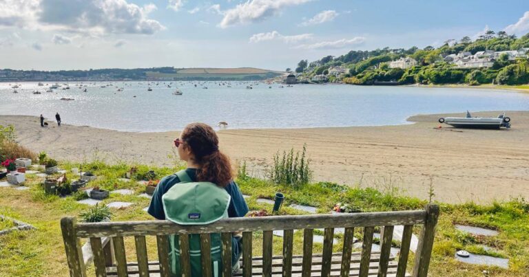 Rebecca Broad sits on a bench overlooking a small estuary beach in Cornwall